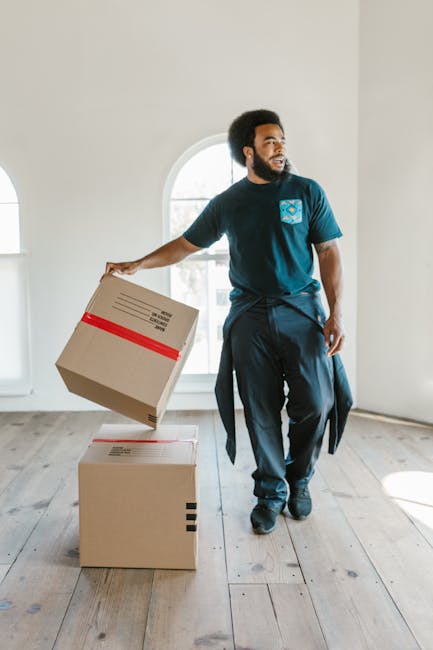 A young woman with curly black hair is sitting on a large cardboard box inside a room with a textured blue wall and wooden floor. She is smiling and resting her chin on her arms, which are folded on t